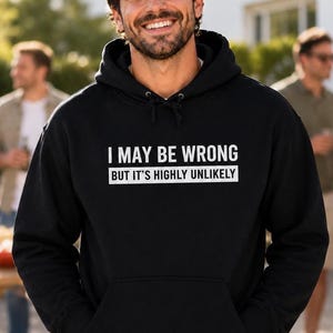 May include: A black hooded sweatshirt featuring white text on the chest that reads I MAY BE WRONG BUT IT'S HIGHLY UNLIKELY. The garment includes a drawstring hood, long sleeves with ribbed cuffs, and a large front kangaroo pocket for storage.