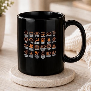 Könnte beinhalten: Ein schwarzer Keramikbecher mit einem Raster aus orangefarbenen Symbolen für Auto-Armaturenbrett-Warnleuchten. Jedes Symbol ist mit humorvollen Textbeschriftungen wie Squishy Doofers, Drunk Robot, Cyclone in Car, Ghosts und Lanky Dalek versehen.