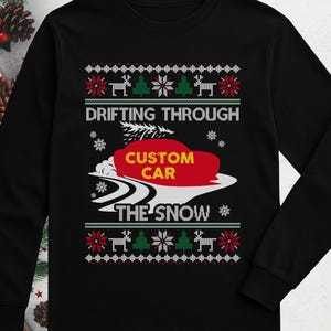 Op de afbeelding: Een zwart shirt met lange mouwen en een afbeelding van een rode auto die door de sneeuw drift, met een kerstboom op het dak en de tekst "DRIFTING THROUGH CUSTOM CAR THE SNOW", omringd door rendieren, bomen en sneeuwvlokken.