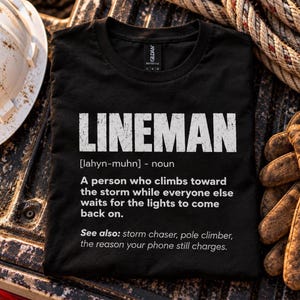 Puede incluir: Una camiseta negra de manga corta con texto blanco que define a un lineman como alguien que escala hacia la tormenta mientras otros esperan a que vuelva la luz. El gráfico incluye la pronunciación fonética y términos relacionados.