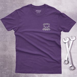 Op de afbeelding: Een paars t-shirt met korte mouwen en een kleine witte afbeelding op de linkerborst. De afbeelding toont een krukas met zuigers en de tekst CRANKY erboven. Het shirt heeft een ronde hals en een label aan de binnenkant van de kraag.