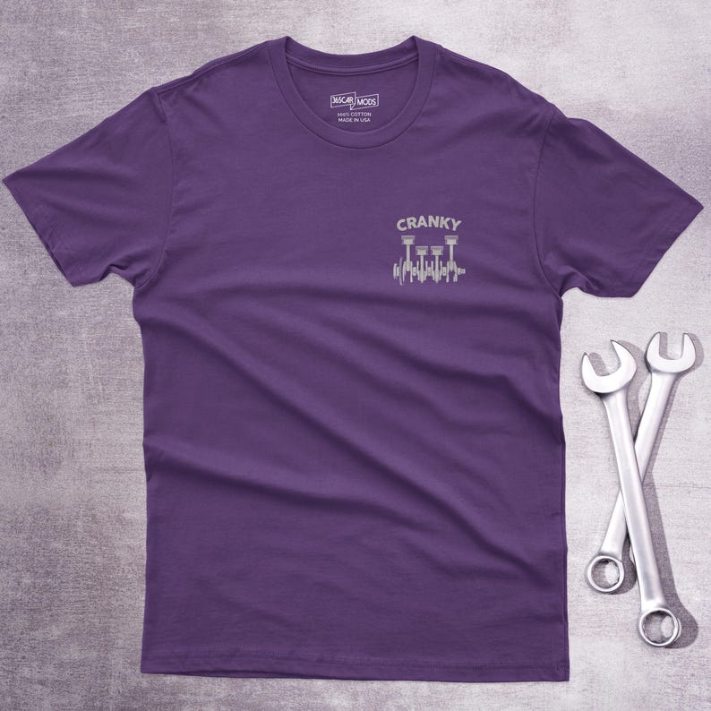 Op de afbeelding: Een paars t-shirt met korte mouwen en een kleine witte afbeelding op de linkerborst. De afbeelding toont een krukas met zuigers en de tekst CRANKY erboven. Het shirt heeft een ronde hals en een label aan de binnenkant van de kraag.