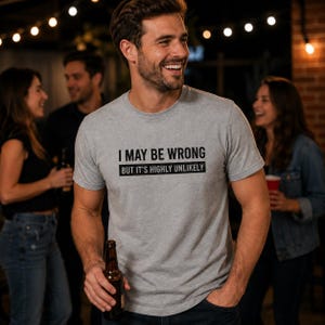 May include: A heather gray short-sleeved crew neck t-shirt featuring the black printed text I MAY BE WRONG BUT IT'S HIGHLY UNLIKELY. The shirt is made of a soft cotton-blend fabric and has a standard fit with a straight hemline and short sleeves.