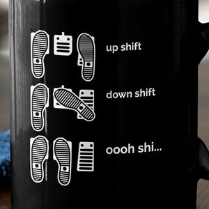 Puede incluir: Una taza de cer&aacute;mica negra con un estampado gr&aacute;fico blanco de tres filas de pedales de coche y huellas. El texto dice up shift, down shift y oooh shi..., ilustrando las posiciones de los pies para conducir con transmisi&oacute;n manual.