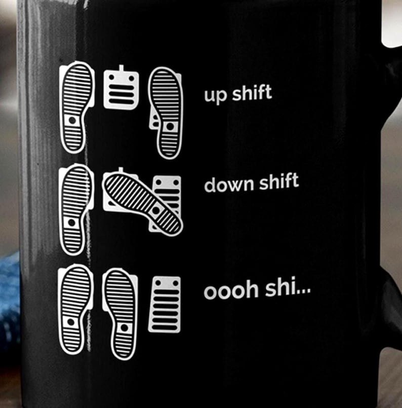 Puede incluir: Una taza de cer&aacute;mica negra con un estampado gr&aacute;fico blanco de tres filas de pedales de coche y huellas. El texto dice up shift, down shift y oooh shi..., ilustrando las posiciones de los pies para conducir con transmisi&oacute;n manual.