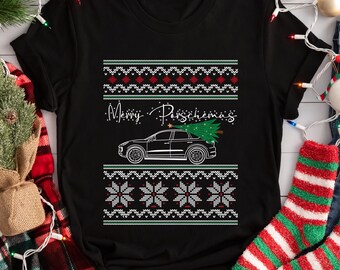 May include: Black t-shirt with a festive design. The shirt features a car carrying a Christmas tree, with the text "Merry Porschemas" above. The design includes a pattern of snowflakes and decorative elements in red, white, and green.