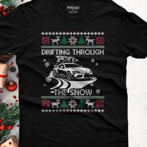 May include: A black short-sleeve t-shirt featuring a graphic print with the text "DRIFTING THROUGH THE SNOW". The design includes a white sports car, snowflakes, and a festive pattern of reindeer and pine trees in red, green, and white colors.