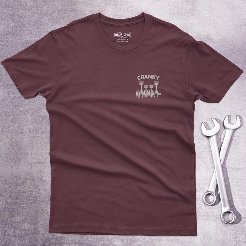 Op de afbeelding: Een bordeauxrood t-shirt met korte mouwen en een witte afbeelding op de linkerborst. De afbeelding toont een krukas met zuigers en de tekst CRANKY erboven. Het label in de nek vermeldt CAR MODS 100% COTTON MADE IN USA.