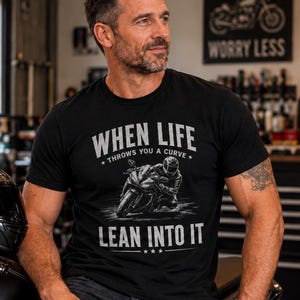 May include: A black short-sleeved t-shirt featuring a graphic of a motorcyclist leaning into a turn. The text reads "WHEN LIFE THROWS YOU A CURVE" above the graphic and "LEAN INTO IT" below, with three small stars centered at the very bottom.