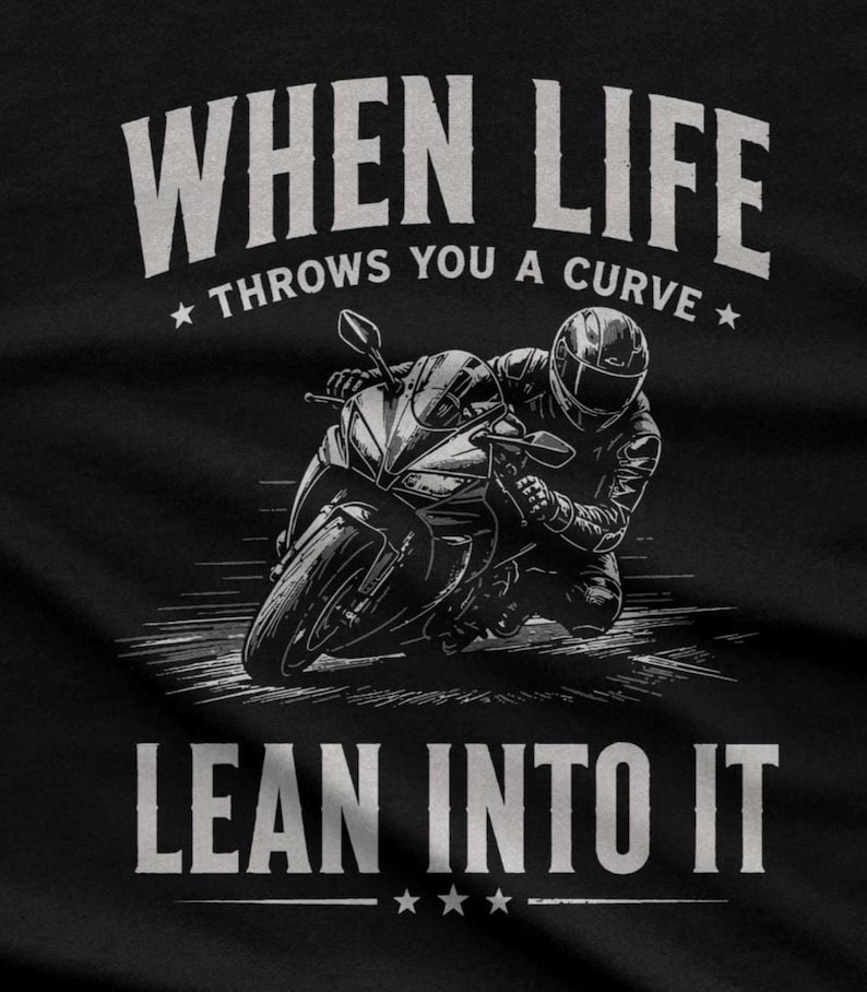 May include: A black t-shirt featuring a white graphic of a motorcyclist leaning into a turn on a sportbike. The text reads "WHEN LIFE THROWS YOU A CURVE" above the motorcycle and "LEAN INTO IT" below, accented with small star icons.