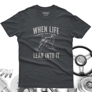 May include: A charcoal gray short-sleeve t-shirt featuring a graphic of a motorcyclist leaning into a turn. The text reads "WHEN LIFE THROWS YOU A CURVE LEAN INTO IT" in a light gray font. The shirt has a crew neck and a standard fit design.