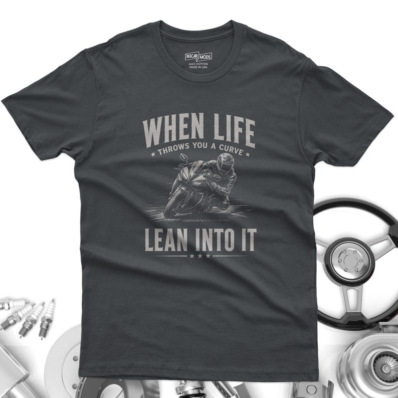 May include: A charcoal gray short-sleeve t-shirt featuring a graphic of a motorcyclist leaning into a turn. The text reads "WHEN LIFE THROWS YOU A CURVE LEAN INTO IT" in a light gray font. The shirt has a crew neck and a standard fit design.