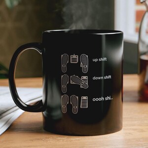 Puede incluir: Una taza de cer&aacute;mica negra con una ilustraci&oacute;n gr&aacute;fica blanca de pedales de coche y pies. El dise&ntilde;o muestra tres filas etiquetadas como up shift, down shift y oooh shi... con las posiciones de los pies sobre los pedales.
