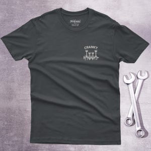 Op de afbeelding: Een donkergrijs t-shirt met korte mouwen en een kleine witte afbeelding op de linkerborst. De afbeelding toont een mechanische krukas met zuigers en de tekst CRANKY erboven. Het label in de nek bevat de tekst OSCAR MODS 100% COTTON.