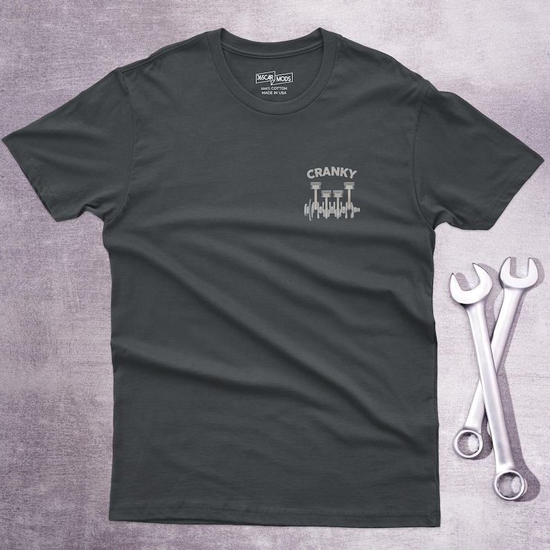 Op de afbeelding: Een donkergrijs t-shirt met korte mouwen en een kleine witte afbeelding op de linkerborst. De afbeelding toont een mechanische krukas met zuigers en de tekst CRANKY erboven. Het label in de nek bevat de tekst OSCAR MODS 100% COTTON.