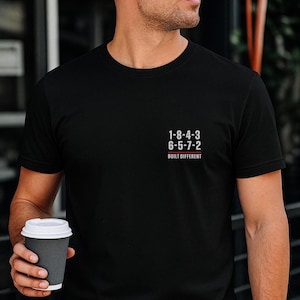 Könnte beinhalten: Ein schwarzes kurzärmeliges T-Shirt mit einer kleinen Grafik auf der linken Brust. Die Grafik zeigt die Zahlen 1-8-4-3 und 6-5-7-2 in Weiß, getrennt durch eine dünne rote horizontale Linie, mit dem Text BUILT DIFFERENT darunter in Weiß.