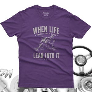 May include: A purple short-sleeve t-shirt featuring a graphic of a motorcyclist leaning into a curve. The text reads "WHEN LIFE THROWS YOU A CURVE LEAN INTO IT" in light gray lettering. The shirt has a crew neckline and a standard fit.