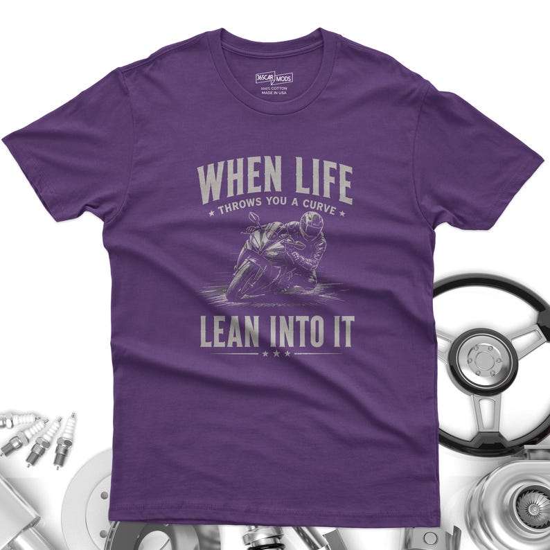 May include: A purple short-sleeve t-shirt featuring a graphic of a motorcyclist leaning into a curve. The text reads "WHEN LIFE THROWS YOU A CURVE LEAN INTO IT" in light gray lettering. The shirt has a crew neckline and a standard fit.