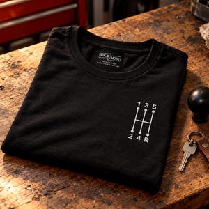 Peut inclure: Un t-shirt noir avec un motif blanc représentant une grille de levier de vitesse manuelle avec les chiffres 1, 3, 5, 2, 4 et R. L'étiquette intérieure du col affiche le texte 365CAR MODS, 100% COTTON, et WASH COLD HANG DRY en blanc.