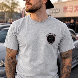 May include: A heather gray short-sleeve t-shirt featuring a circular graphic on the left chest. The graphic displays a manual transmission gear shift pattern with the text "ENDANGERED SPECIES" arched around the top and bottom of the circle.