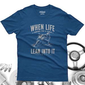 May include: A blue short-sleeved t-shirt featuring a graphic of a motorcyclist leaning into a turn. The text reads "WHEN LIFE THROWS YOU A CURVE LEAN INTO IT" in light gray lettering. The shirt has a crew neckline and a standard fit design.