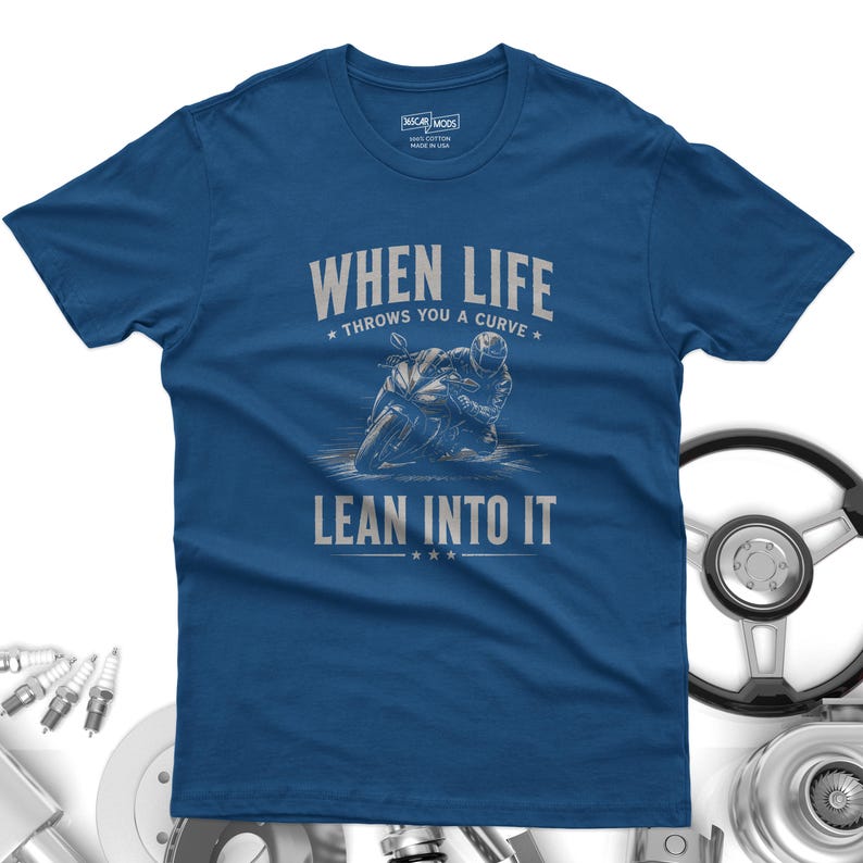 May include: A blue short-sleeved t-shirt featuring a graphic of a motorcyclist leaning into a turn. The text reads "WHEN LIFE THROWS YOU A CURVE LEAN INTO IT" in light gray lettering. The shirt has a crew neckline and a standard fit design.