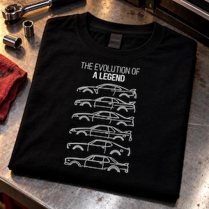 May include: A black t-shirt featuring a white graphic print of six car silhouettes stacked vertically. Above the cars, the text reads THE EVOLUTION OF A LEGEND in white capital letters. The shirt has a crew neckline and short sleeves.