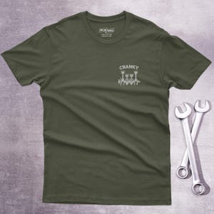 Op de afbeelding: Een olijfgroen t-shirt met korte mouwen en een kleine witte afbeelding op de linkerborst. De afbeelding toont een krukas met zuigers en de tekst CRANKY erboven. Het shirt heeft een ronde hals en een label in de kraag.