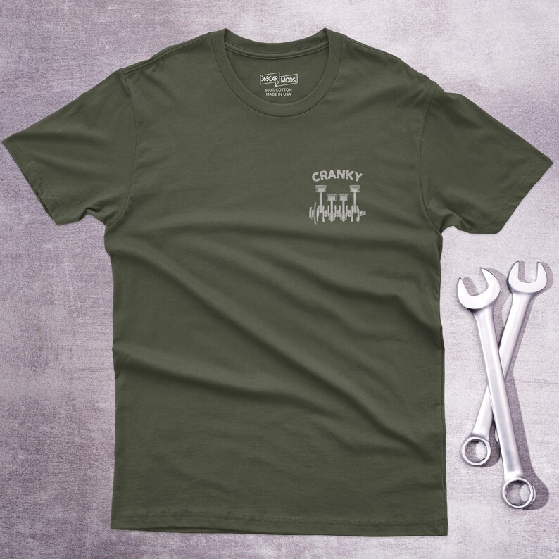 Op de afbeelding: Een olijfgroen t-shirt met korte mouwen en een kleine witte afbeelding op de linkerborst. De afbeelding toont een krukas met zuigers en de tekst CRANKY erboven. Het shirt heeft een ronde hals en een label in de kraag.