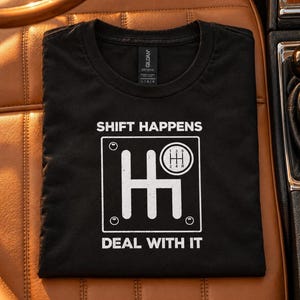May include: A black t-shirt featuring a white graphic of a manual transmission gear shift pattern. The text SHIFT HAPPENS is printed above the graphic, and the text DEAL WITH IT is printed below the graphic in a bold, white, sans-serif font.