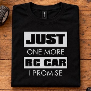 Könnte beinhalten: Ein schwarzes T-Shirt mit Rundhalsausschnitt und dem weißen Schriftzug JUST ONE MORE RC CAR I PROMISE in einer fetten Blockschrift. Das T-Shirt hat ein Gildan-Markenetikett am inneren Halsausschnitt und liegt auf einer flachen Holzoberfläche.