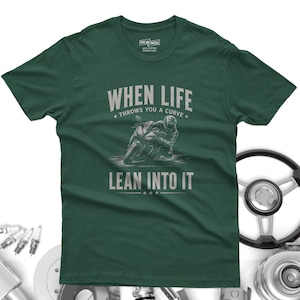 May include: A dark green short-sleeve t-shirt featuring a graphic of a motorcyclist leaning into a curve. The text reads "WHEN LIFE THROWS YOU A CURVE LEAN INTO IT" in a light gray font. The shirt has a crew neckline and a standard fit.