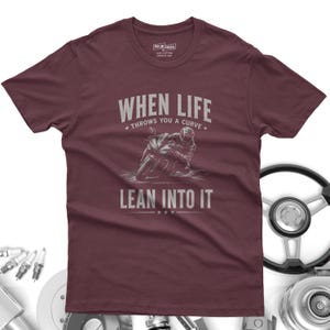 May include: A maroon short-sleeve t-shirt featuring a graphic of a motorcyclist leaning into a curve. The text reads "WHEN LIFE THROWS YOU A CURVE LEAN INTO IT" in a light gray font. The shirt has a crew neckline and a label printed on the inside neck.