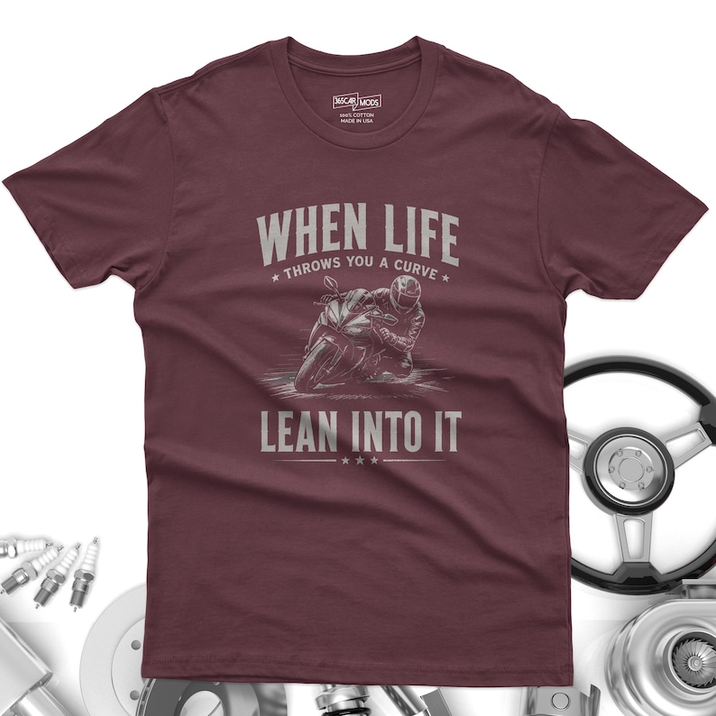 May include: A maroon short-sleeve t-shirt featuring a graphic of a motorcyclist leaning into a curve. The text reads "WHEN LIFE THROWS YOU A CURVE LEAN INTO IT" in a light gray font. The shirt has a crew neckline and a label printed on the inside neck.