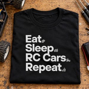 Könnte beinhalten: Ein schwarzes T-Shirt mit kurzen Ärmeln und dem weißen Schriftzug Eat, Sleep, RC Cars, Repeat. Jede Textzeile wird von einer kleinen weißen Grafik eines ferngesteuerten Fahrzeugs begleitet, darunter ein Auto, ein Truck und ein Buggy.