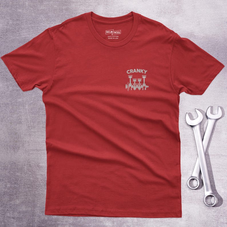 Op de afbeelding: Een rood t-shirt met korte mouwen en een kleine afbeelding op de linkerborst. De afbeelding toont een zilverkleurige illustratie van een krukas en zuigers met de tekst CRANKY erboven gedrukt. Het t-shirt heeft een ronde hals.