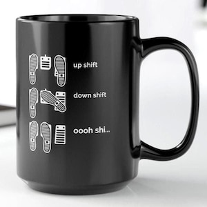 Puede incluir: Una taza de cer&aacute;mica negra con un estampado gr&aacute;fico blanco de configuraciones de pedales de coche. El dise&ntilde;o muestra tres filas de posiciones de pies con el texto up shift, down shift y oooh shi... junto a ilustraciones de pedales.