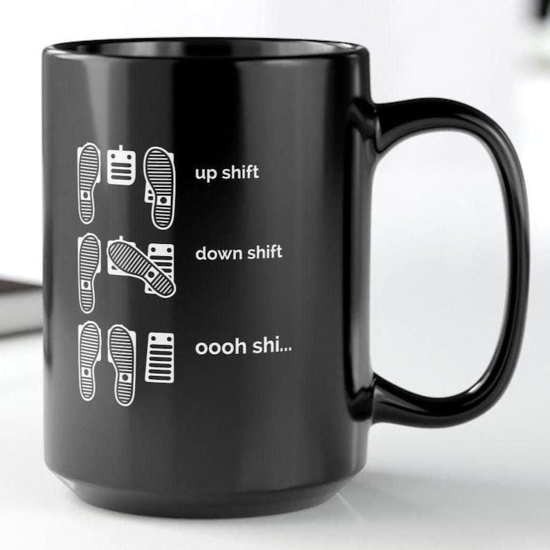 Puede incluir: Una taza de cer&aacute;mica negra con un estampado gr&aacute;fico blanco de configuraciones de pedales de coche. El dise&ntilde;o muestra tres filas de posiciones de pies con el texto up shift, down shift y oooh shi... junto a ilustraciones de pedales.