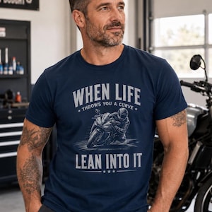 May include: A navy blue short-sleeve t-shirt featuring a graphic of a motorcyclist leaning into a turn. The text reads "WHEN LIFE THROWS YOU A CURVE LEAN INTO IT" in a light gray font. The design includes small star accents above and below the text.