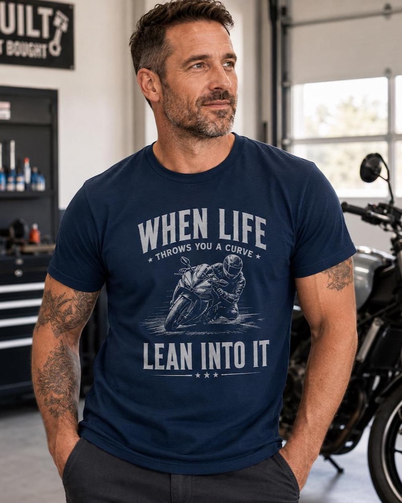 May include: A navy blue short-sleeve t-shirt featuring a graphic of a motorcyclist leaning into a turn. The text reads "WHEN LIFE THROWS YOU A CURVE LEAN INTO IT" in a light gray font. The design includes small star accents above and below the text.