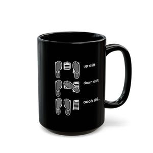 Puede incluir: Una taza de cer&aacute;mica negra con un estampado gr&aacute;fico blanco de tres filas de pedales de coche y huellas de zapatos. El texto dice up shift, down shift y oooh shi... junto a las posiciones de los pedales, ilustrando el cambio de marchas manual.