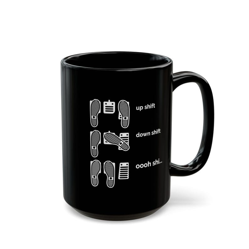 Puede incluir: Una taza de cer&aacute;mica negra con un estampado gr&aacute;fico blanco de tres filas de pedales de coche y huellas de zapatos. El texto dice up shift, down shift y oooh shi... junto a las posiciones de los pedales, ilustrando el cambio de marchas manual.