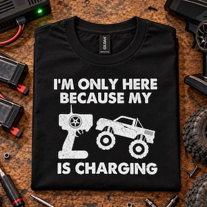 Puede incluir: Una camiseta negra con texto blanco desgastado que dice I'M ONLY HERE BECAUSE MY IS CHARGING, acompañada de una ilustración gráfica de un control remoto y un camión monstruo colocada entre las palabras MY e IS en la prenda.