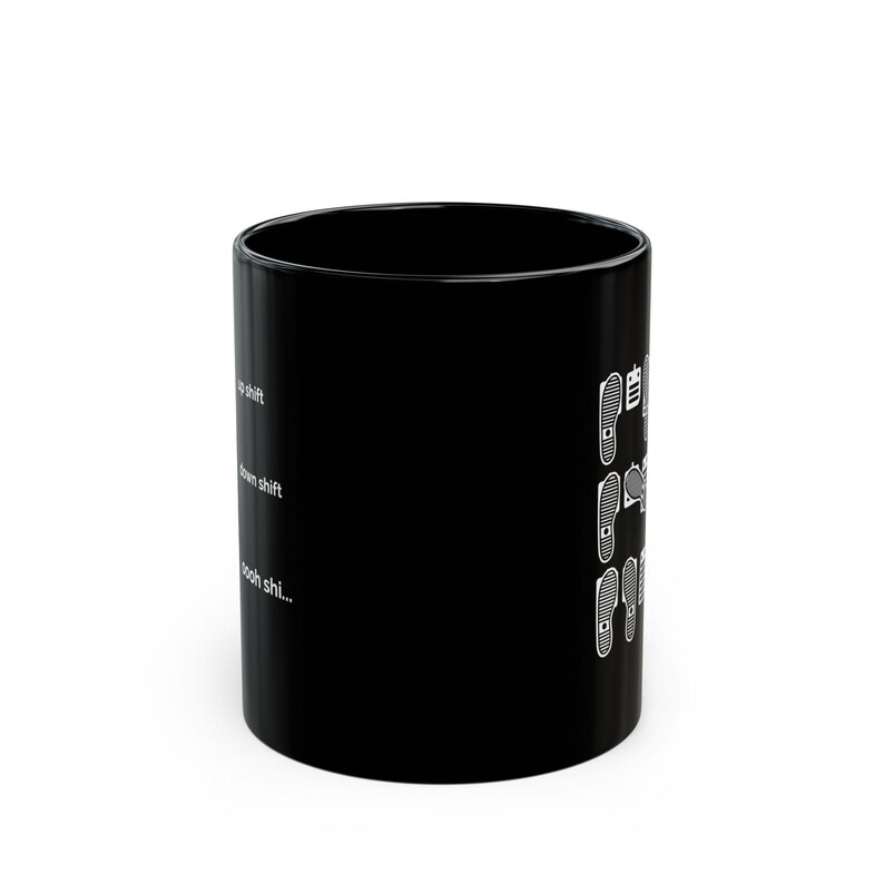 Puede incluir: Una taza de cer&aacute;mica negra con gr&aacute;ficos blancos de suelas de zapatos sobre pedales de coche. El texto dice up shift, down shift y oooh shi... dispuesto verticalmente junto a las ilustraciones de los pedales, representando una transmisi&oacute;n manual.