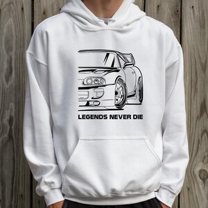 Puede incluir: Sudadera con capucha blanca con un gr&aacute;fico negro de un coche deportivo y el texto "LEGENDS NEVER DIE" debajo. El dise&ntilde;o del coche es un dibujo lineal detallado de la mitad delantera de un veh&iacute;culo.