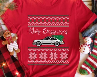 May include: Red t-shirt with a festive design. The shirt features a white car with a green Christmas tree on top, and the text "Merry Classicmas". The design includes white snowflakes and green and white patterns.