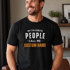 May include: A black short-sleeved t-shirt featuring the white text MY FAVORITE PEOPLE CALL ME and the orange text CUSTOM NAME printed on the chest. The shirt has a crew neckline and a standard fit, displaying a clear graphic design on the front.