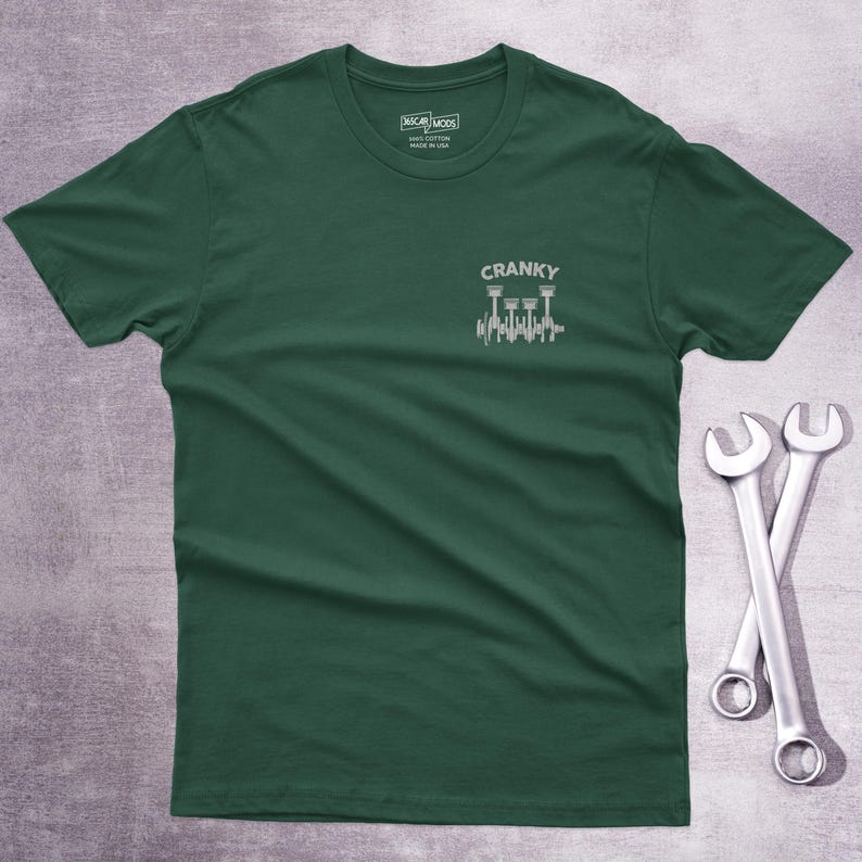 Op de afbeelding: Een donkergroen T-shirt met korte mouwen en een kleine witte afbeelding op de linkerborst. De afbeelding toont een krukas met zuigers en de tekst CRANKY erboven. Het label in de nek vermeldt OSCAR MODS 100% COTTON MADE IN USA.