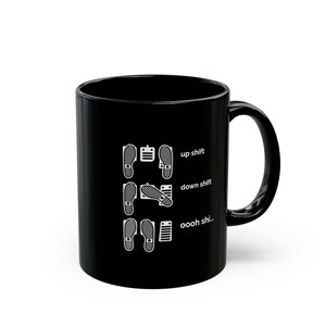 Puede incluir: Una taza de cer&aacute;mica negra con un gr&aacute;fico blanco de tres filas de pedales de coche y posiciones de pies. El texto dice up shift, down shift y oooh shi... junto a las configuraciones de pedales para un veh&iacute;culo de transmisi&oacute;n manual.