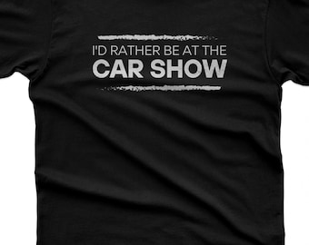 Pode incluir: Uma t-shirt preta de manga curta com o texto branco "I'D RATHER BE AT THE CAR SHOW" centrado no peito. O texto é emoldurado por duas linhas brancas horizontais com aspeto desgastado. A etiqueta interior mostra a marca 365CAR MODS.
